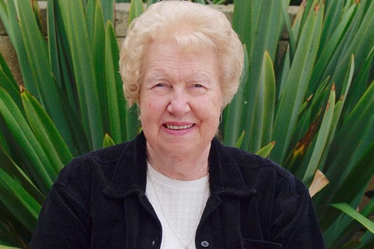 Portrait of Dolores Cannon, founder of QHHT®, smiling warmly in front of green foliage, known for her work in past life regression and soul exploration.